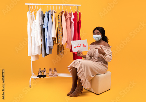 Young black woman pointing at SALE sign, sitting near rack with clothes, wearing face mask, advertising clothing store