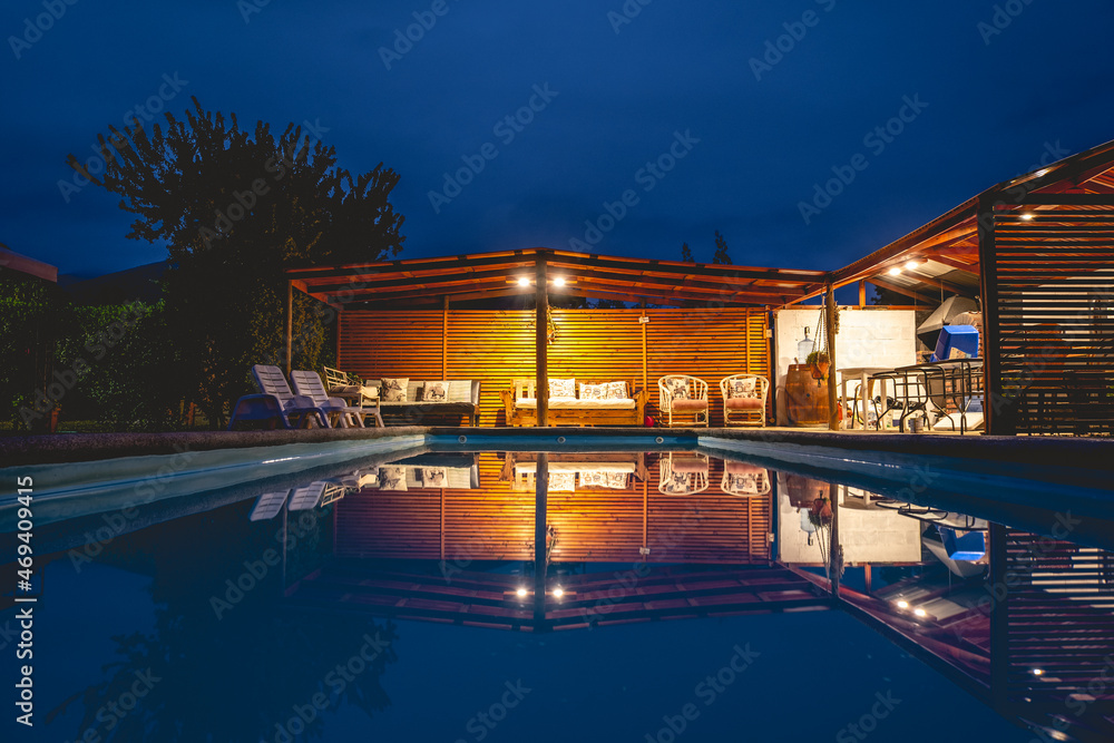 Obraz premium View of a big beautiful pool and wooden pavilion with barbecue in the countryside with beautiful lights and the blue sky in the night