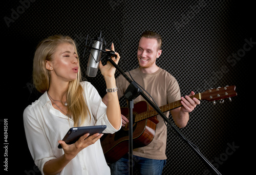 Young female singing with tablet computer in her hand. While the man playing an acoustic guitar in front of black soundproofing walls. Musicians producing music in professional recording studio. © chadchai
