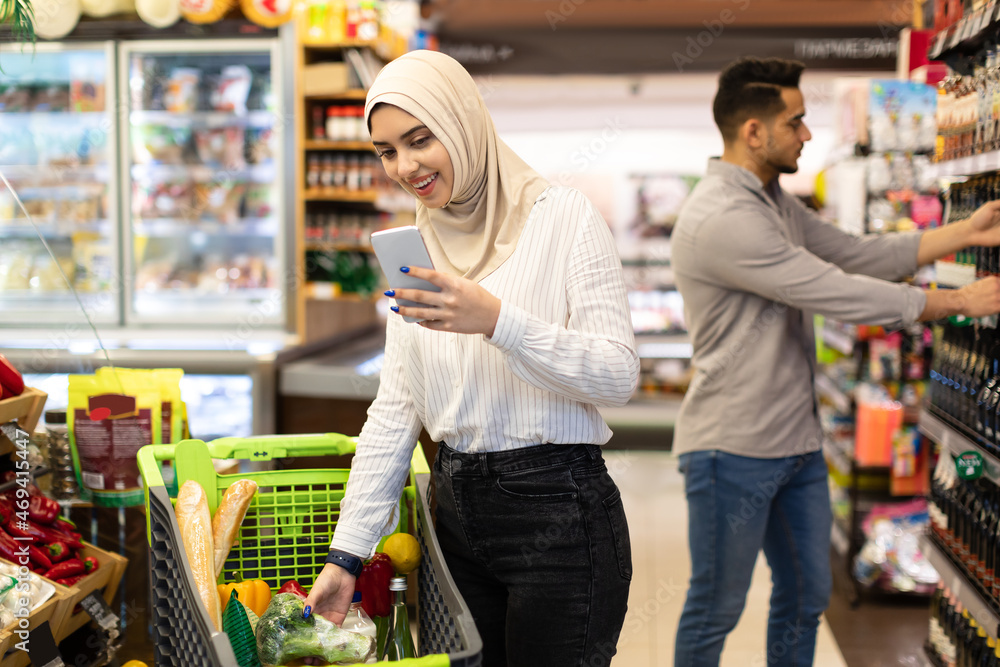 Islamic Lady Doing Grocery Shopping Using Mobile Phone In Supermarket