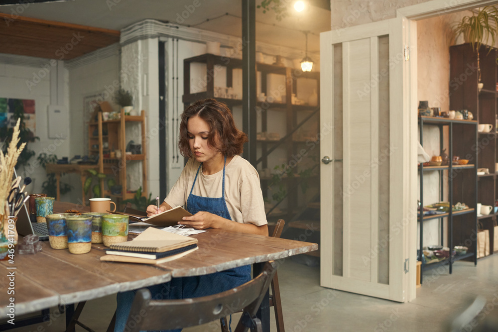 Young female ceramist in workwear making notes while sitting by table in front of laptop in her workshop Young female ceramist in workwear making notes while sitting by table in front of laptop in her workshop