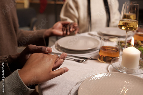 Young African man holding his wife and daughter by hands while sitting by served festive table and praying before dinner