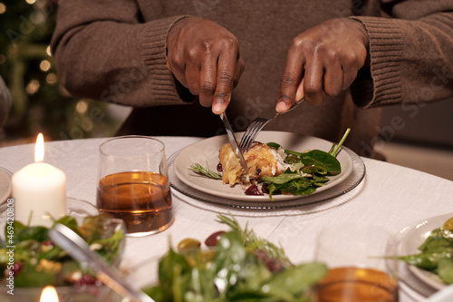 Hands of young man with knife and fork cutting piece of chicken on plate while having festive dinner by served table