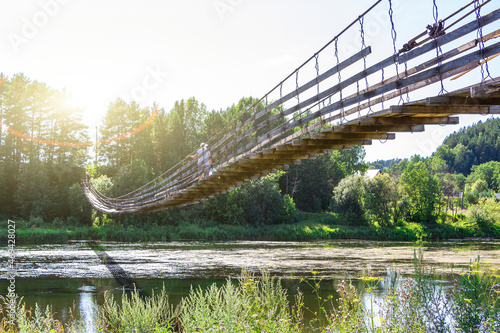 Woman with dog walks along old wooden hanging bridge on river in summer sunny day. Natural landscape. Dog on bridge