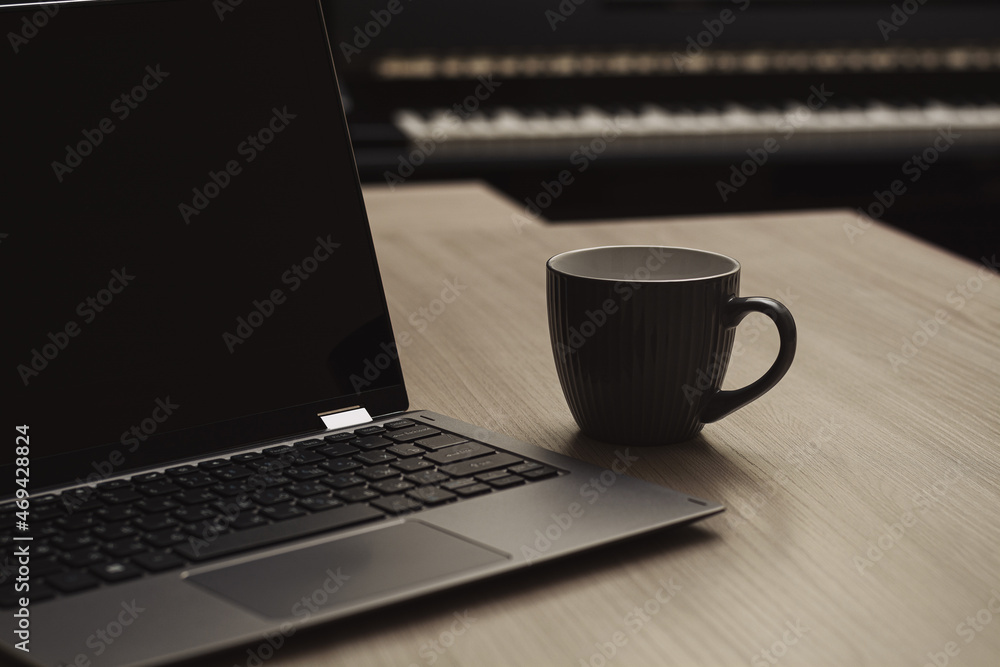 A gray cup of coffee next to a laptop on a wooden table against the background of a piano. Home Office. Horizontal view. Selective focus. A gray cup of coffee next to a laptop on a wooden table against the background of a piano. Home Office. Horizontal view. Selective focus.