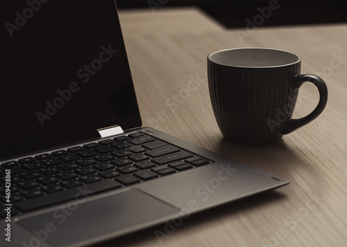 Close-up gray cup of coffee next to a laptop on a wooden table. Home Office. Horizontal view. Selective focus.
