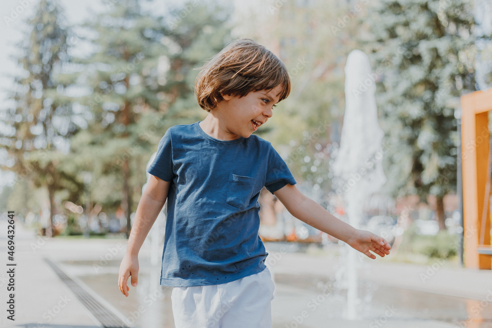Naklejka premium little boy is running in the street and playing with the water jets of a fountain spouting from the ground. child in blue T-shirt and white shorts is fooling around outdoors. lifestyle. space for text