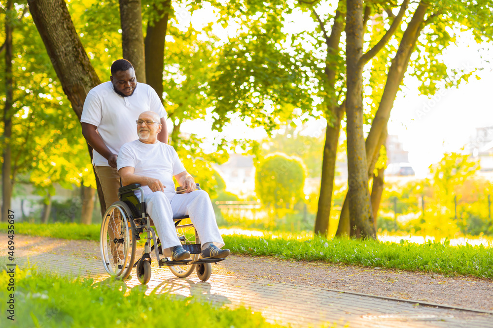 Caregiver and old man in a wheelchair. Professional nurse and patient walking outdoor in the park at sunset. Assistance, rehabilitation and health care. Caregiver and old man in a wheelchair. Professional nurse and patient walking outdoor in the park at sunset. Assistance, rehabilitation and health care.