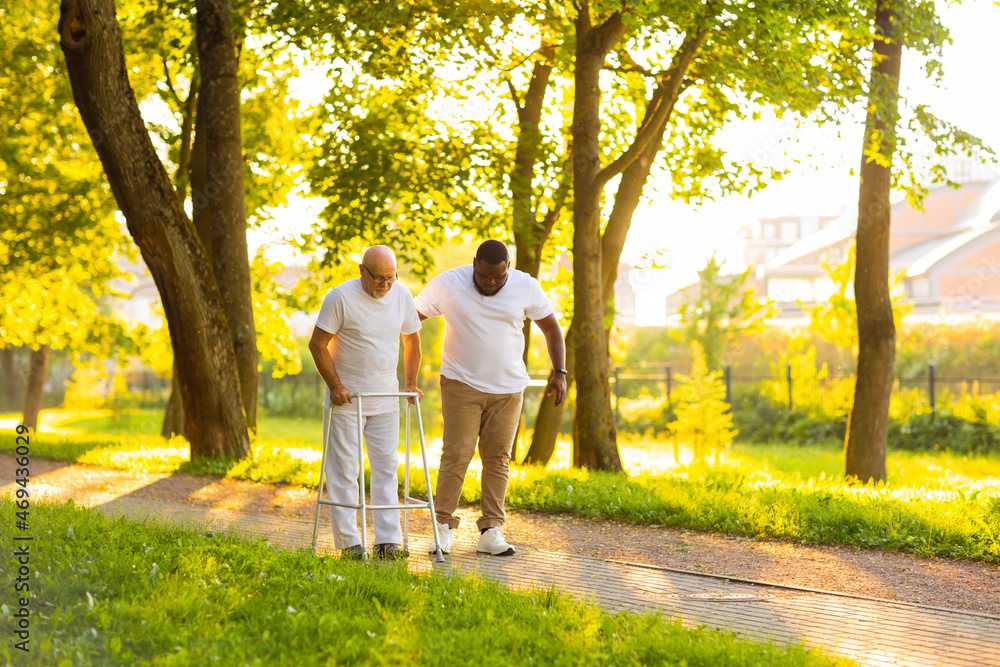 Obraz premium Caregiver is teaching old man to walk with walker. Professional nurse and patient walking outdoor at sunset. Assistance, rehabilitation and health care.