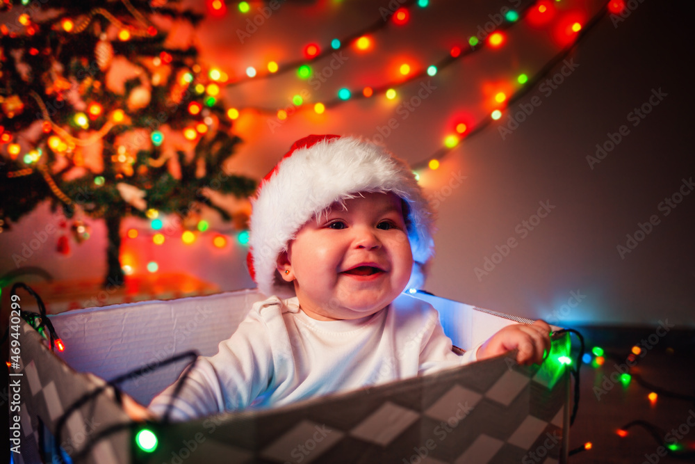 Christmas. Portrait of a happy baby in a Christmas hat sitting in a gift box. A garland lights up in the background. The concept of winter Christmas holidays Christmas. Portrait of a happy baby in a Christmas hat sitting in a gift box. A garland lights up in the background. The concept of winter Christmas holidays