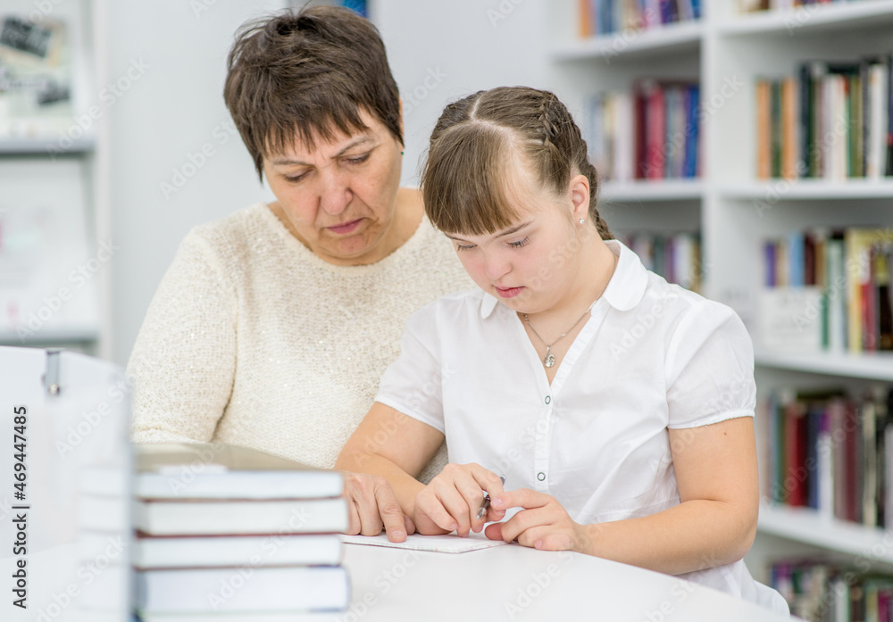 Senior woman helps to girl with Downs syndrome doing homework at a school. Education for disabled children concept Senior woman helps to girl with Downs syndrome doing homework at a school. Education for disabled children concept
