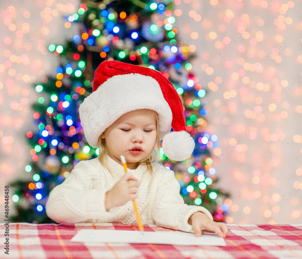 Baby girl wearing red santa hat writes letter to Santa Claus. Festive background with christmas tree Baby girl wearing red santa hat writes letter to Santa Claus. Festive background with christmas tree