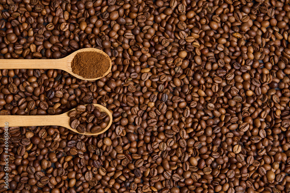 Obraz premium Top view of wooden spoons with ground coffee and roasted coffee beans on the background of scattered fresh roasted coffee grains with copy space for advertisement. Flat lay composition