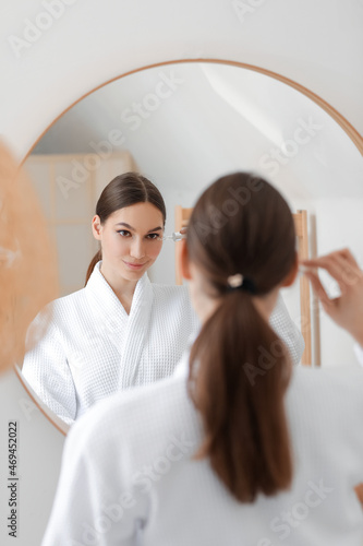 Young woman applying natural essential oil with pipette on her face near mirror