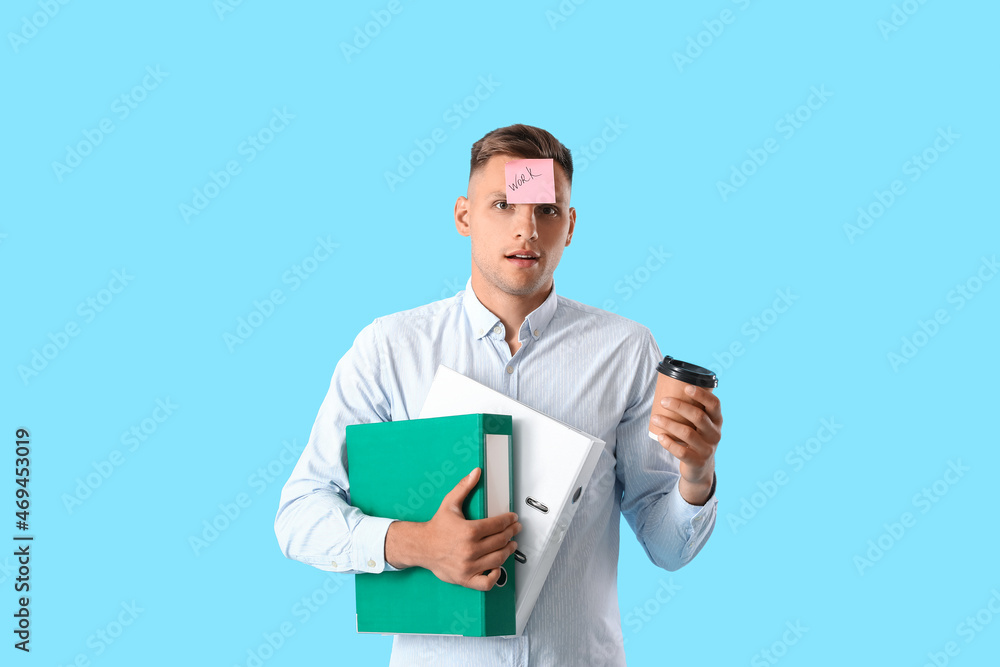 Young man with folders, sticky note with word WORK on his forehead and coffee against color background Young man with folders, sticky note with word WORK on his forehead and coffee against color background
