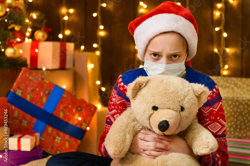 Child girl as Santa helper posing in new year decoration. Wearing a protective face mask against viruses, coronaviruses. Holiday lights and lots of gifts, an elegant Christmas tree with toys.