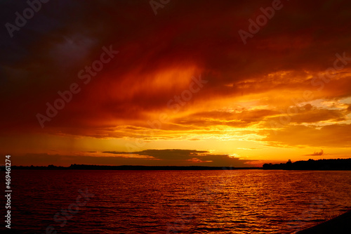 Morning fiery orange sunrise over river. Bloody red cloudy sunset above lake. Reflective water surface waves under clouds at dusk in summer. Evening cloudscape in twilight. Ruddy horizon sky and river