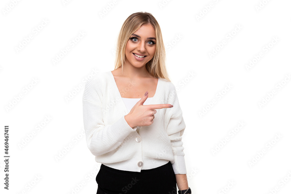 Photo of a beautiful pretty amazing positive smiling happy young blonde woman with makeup in a stylish white knitted sweater and black pants shows finger to the right isolated on a white background Photo of a beautiful pretty amazing positive smiling happy young blonde woman with makeup in a stylish white knitted sweater and black pants shows finger to the right isolated on a white background