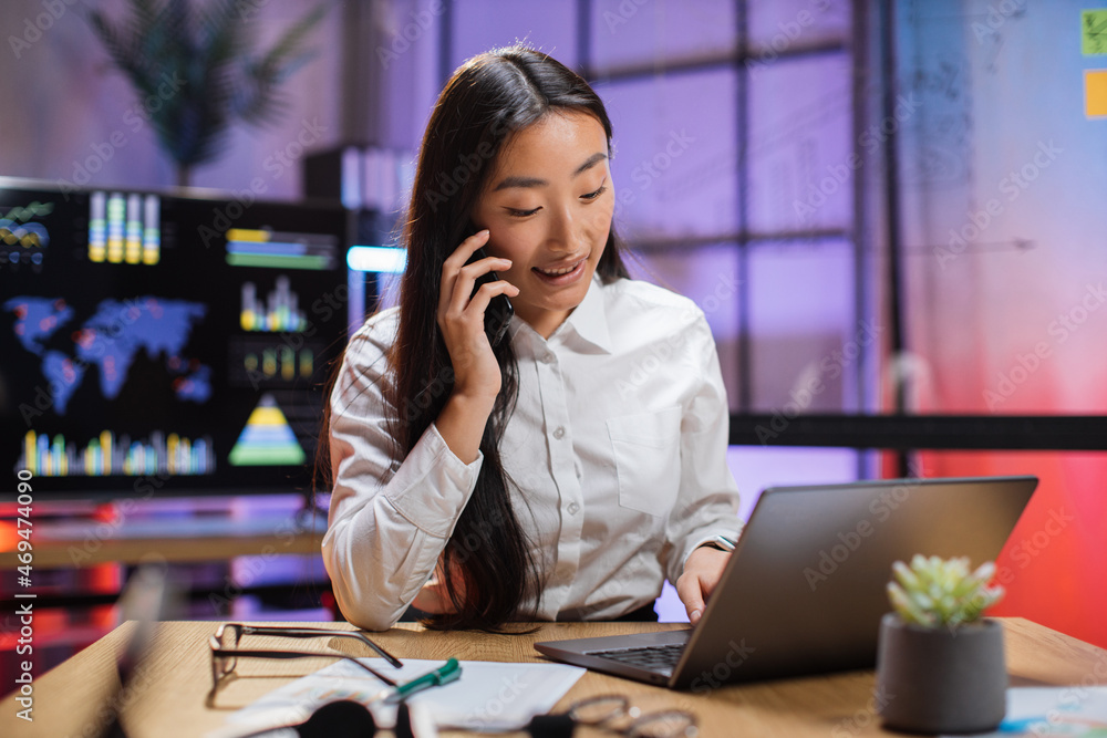 Charming asian woman talking on mobile and typing on laptop at modern office room. Female company member in formal clothes sitting at desk and working. Charming asian woman talking on mobile and typing on laptop at modern office room. Female company member in formal clothes sitting at desk and working.