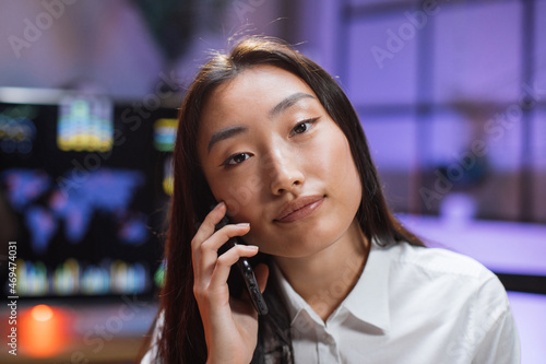 Asian woman with dark hair talking on smartphone while sitting at table. Business lady in white shirt having mobile conversation during work at office.