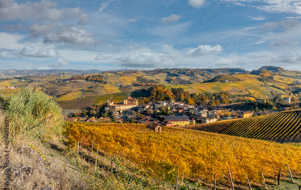 Landscape on the hills of the Barolo area, Langhe, Italy, with Barolo Village and Castle, fine vineyards of Nebbiolo grapes with autumn colors on blue sky with cloud