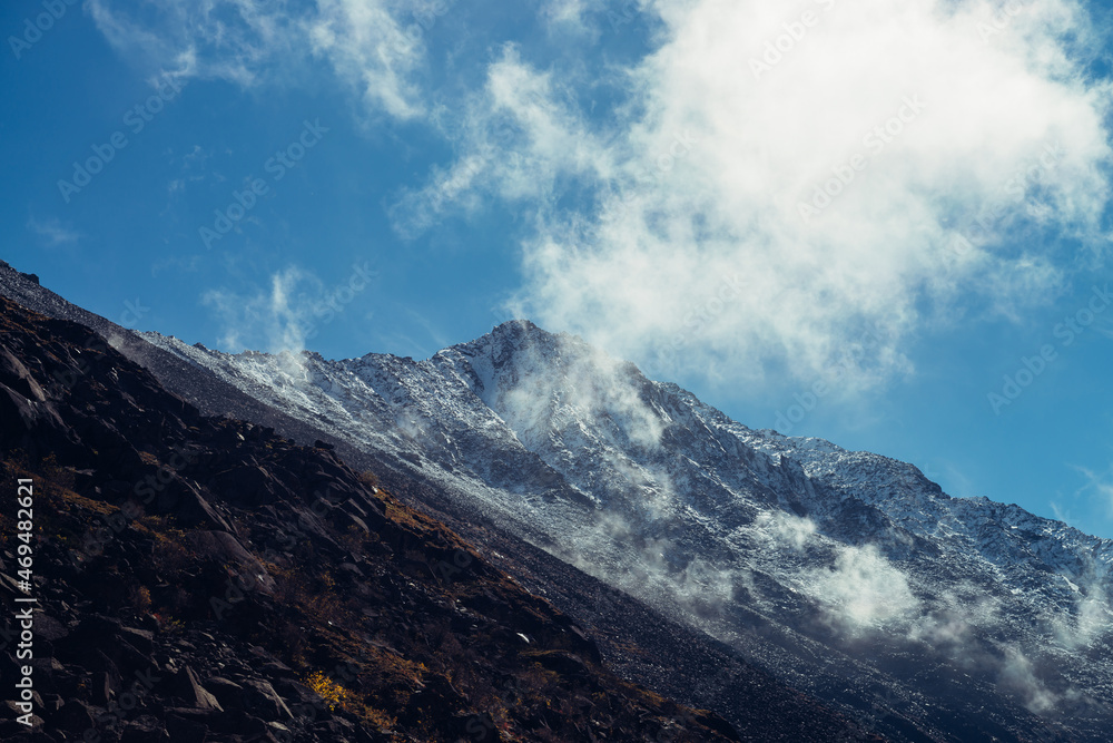 Atmospheric mountain landscape with high black rock peak with snow in low clouds. Beautiful alpine scenery with great mountain with peaked top and low cirrus clouds. Awesome mountain top among clouds.