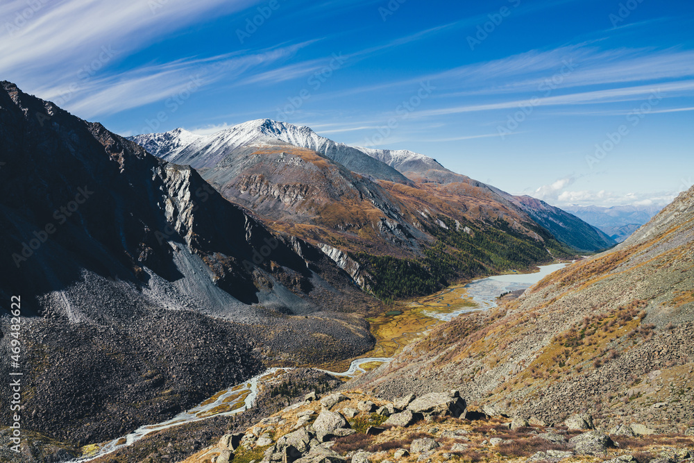 Picturesque highland landscape with mountain river and lake in valley among high mountains with snow in autumn colors in sunshine. Awesome alpine view to motley mountain valley in sunlight in fall. Picturesque highland landscape with mountain river and lake in valley among high mountains with snow in autumn colors in sunshine. Awesome alpine view to motley mountain valley in sunlight in fall.