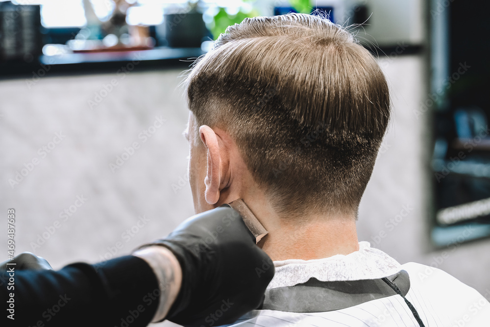 Visit to the barbershop. Hairdresser, barber shaves client's temples,sides with hair clipper. Young stylish man makes fashionable haircut.Customer sitting in dark men's beauty salon covered with cape Visit to the barbershop. Hairdresser, barber shaves client's temples,sides with hair clipper. Young stylish man makes fashionable haircut.Customer sitting in dark men's beauty salon covered with cape