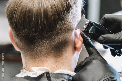 Visit to the barbershop. Hairdresser, barber shaves client's temples,sides with hair clipper. Young stylish man makes fashionable haircut.Customer sitting in dark men's beauty salon covered with cape