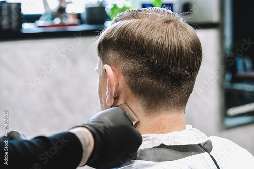 Visit to the barbershop. Hairdresser, barber shaves client's temples,sides with hair clipper. Young stylish man makes fashionable haircut.Customer sitting in dark men's beauty salon covered with cape