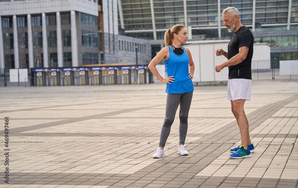 Full length shot of two sportive friends, middle aged man and woman in sportswear talking while standing outdoors, having morning workout