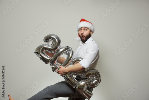 An astonished man with a beard in a red velvet Santa hat is holding silver balloons in the shape of 2022. New year party.
