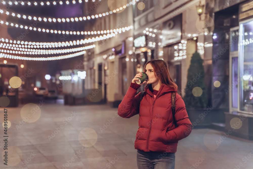 Fototapeta premium Girl in night Christmas city enjoy coffee and atmosphere. Young woman outdoors in winter. Lights on New Years Eve