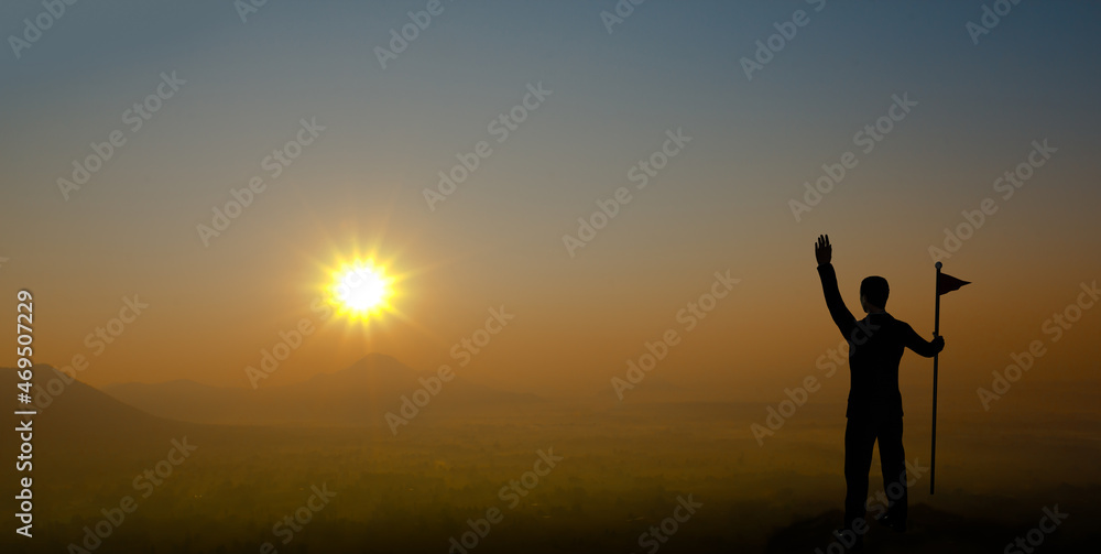 Businessman holding flag and raising hand on the mountain with sunset. Silhouette of male successful entrepreneur looking at the sunset.