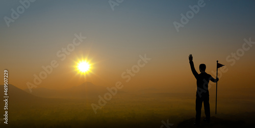 Businessman holding flag and raising hand on the mountain with sunset. Silhouette of male successful entrepreneur looking at the sunset.