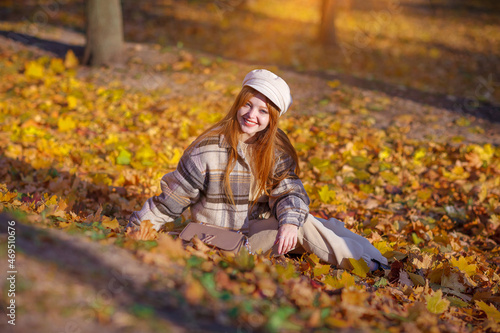 A beautifyl girl with long red hair walks in the park, around the yellow leaves of trees. The woman is dressed in a cap and a warm jacket in cage. Joy, autumn, sunny day, vacation concept.