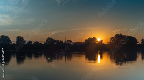 Summer sunset over the river. Trees and clouds reflected in the water. Orange ball of sun hiding behind the forest. The beauty of nature. © VeNN