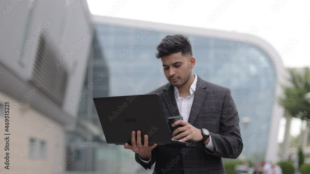 Young male asian indian economist using laptop and drinking coffee to go, standing outside city street. Smiling hindu man posing on camera while working on laptop outdoors office Young male asian indian economist using laptop and drinking coffee to go, standing outside city street. Smiling hindu man posing on camera while working on laptop outdoors office