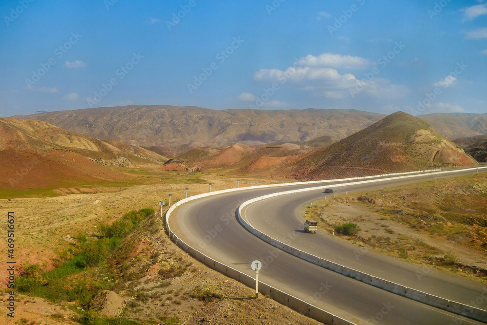 Fototapeta premium Highway with racing cars in background of hilly valley. Mountains are visible in distance. Shot in Surkhandarya region in Uzbekistan near mountains of Gissar ridge