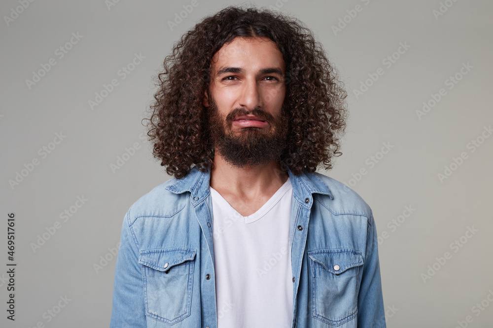 Indoor shot of young bearded male with curly long hair wears denim blue shirt looking directly into camera with serious facial expression. isolated over grey background