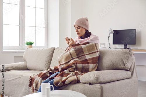 Sick woman who has high temperature due to bad cold or seasonal flu sitting on sofa at home. Sad lady with fever wearing warm hat and plaid looking at thermometer in hand with worried face expression © Studio Romantic