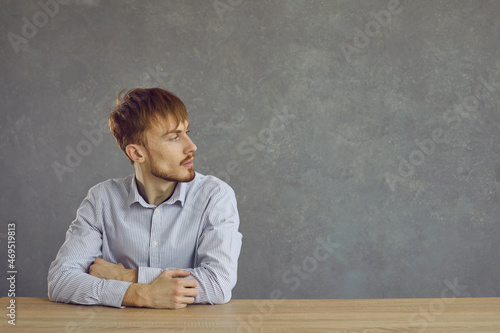 Seated casual young man serious looking to the side against gray background, copy space. For advertising and text. Bearded man looks to the side while sitting at the table.