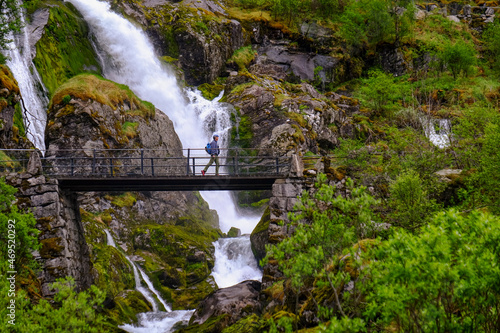 A man on a pedestrian bridge crosses one of many waterfalls originating at the Briksdal glacier, Stryn, Vestland, Norway, Scandinavia photo