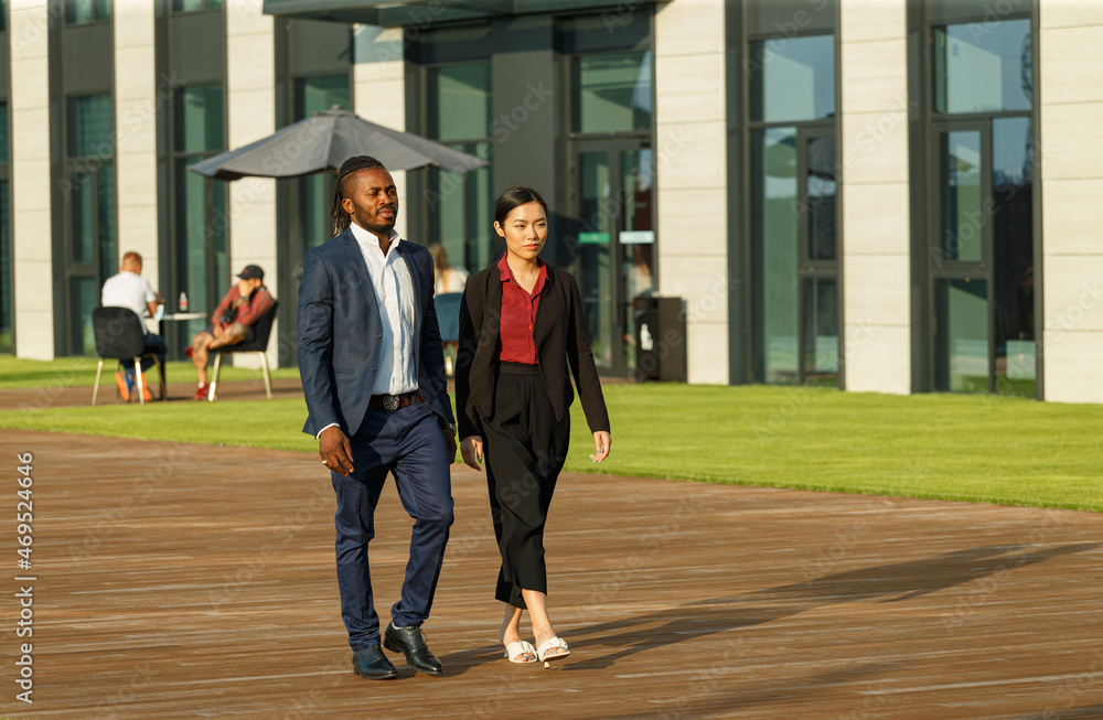 Business partners African American man in formal navy suit and young Asian brunette long-haired woman in red shirt stroll near office