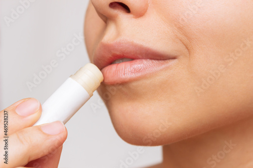 Fotografiet Cropped shot of a young caucasian beautiful woman applying a hygienic lipstick on her lips on a gray background