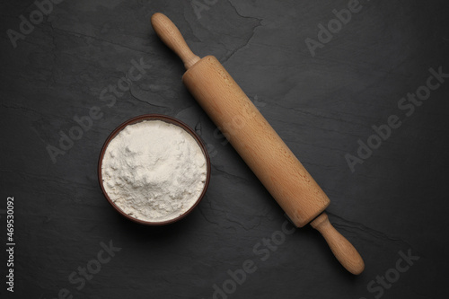 Wooden rolling pin and bowl with flour on black background, flat lay