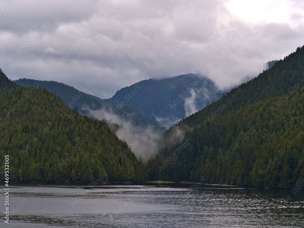 Wilderness landscape with cloud rising from tree-covered mountains at the shore of Tolmnie Channel, part of the Inside Passage, BC, Canada.