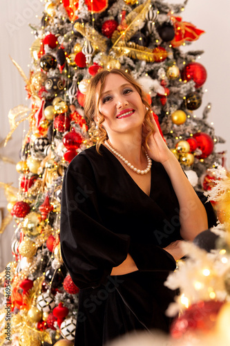 A woman in a black evening dress stands against the background of a Christmas tree. Dress is a Christmas. Party look