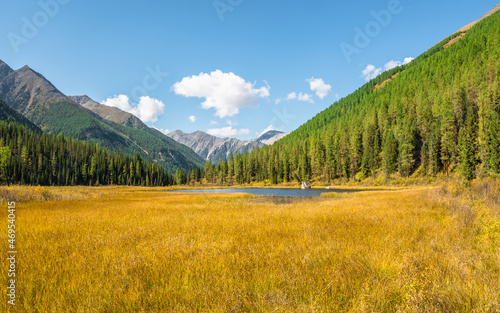 Panoramic view of sunny swampy autumn plateau. Awesome highland scenery with beautiful glacial lake among sunlit hills and rocks against mountain range.