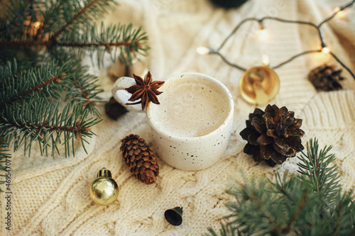 Warm coffee in stylish cup with anise star, fir branches, ornaments, pine cones and warm lights on cozy knitted background. Atmospheric christmas time and hygge winter home
